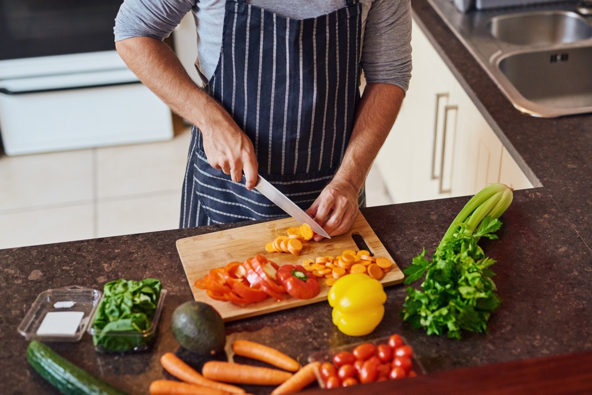 Hands, man and cutting with vegetables for health, nutrition or vegan diet for wellness. Male person, groceries and wood board in kitchen for cooking with food and ingredients to prepare home dinner