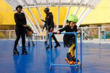 Little girl wearing a neon green helmet, black dress, and yellow tights, holding on to a skate assist on a roller rink